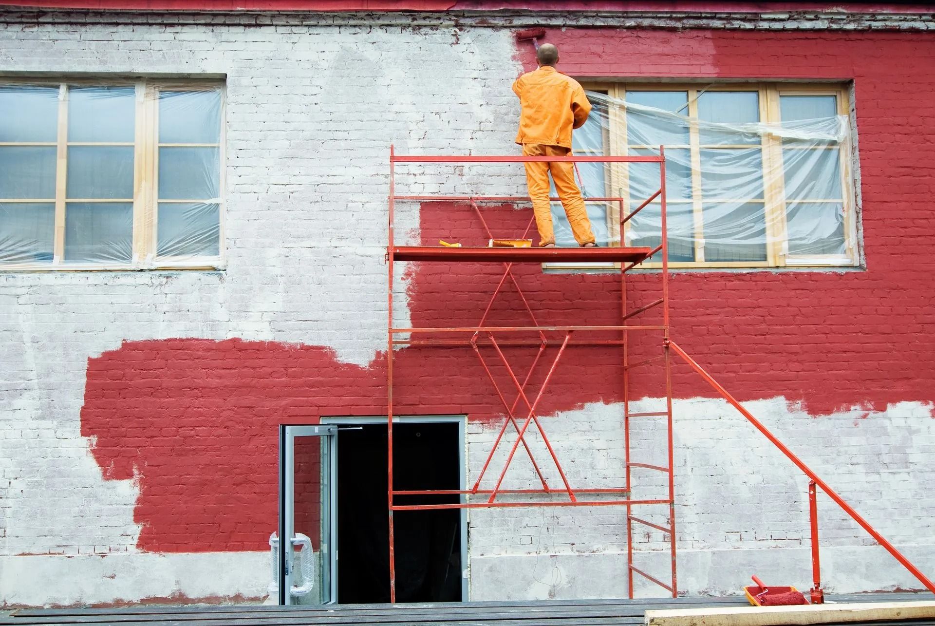 A painter in an orange jumpsuit on scaffolding paints a brick building red; windows are visible.