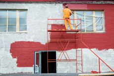 A painter in an orange jumpsuit on scaffolding paints a brick building red; windows are visible.