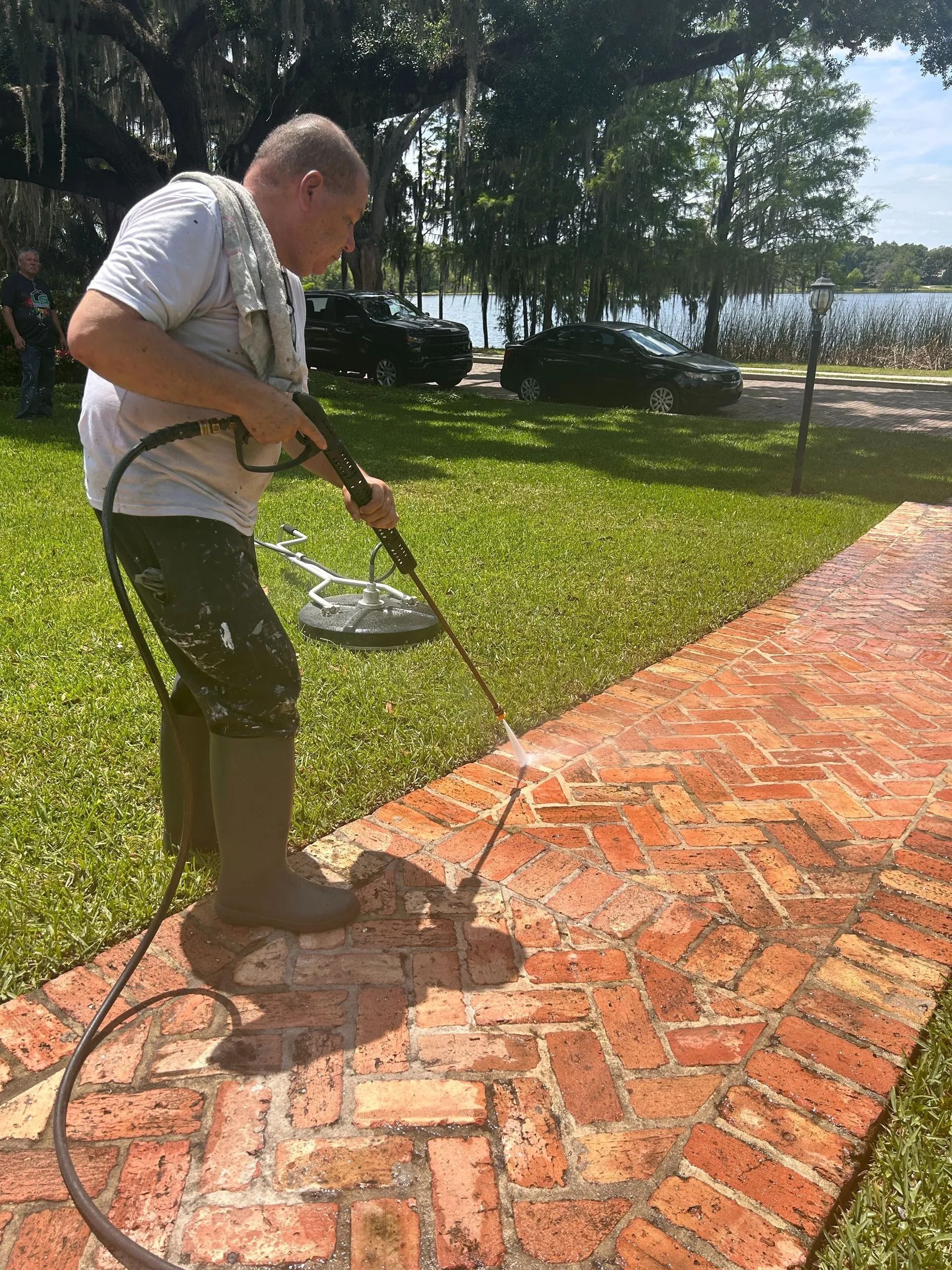 Man power washing a brick walkway with a pressure washer in a yard, near a lake.