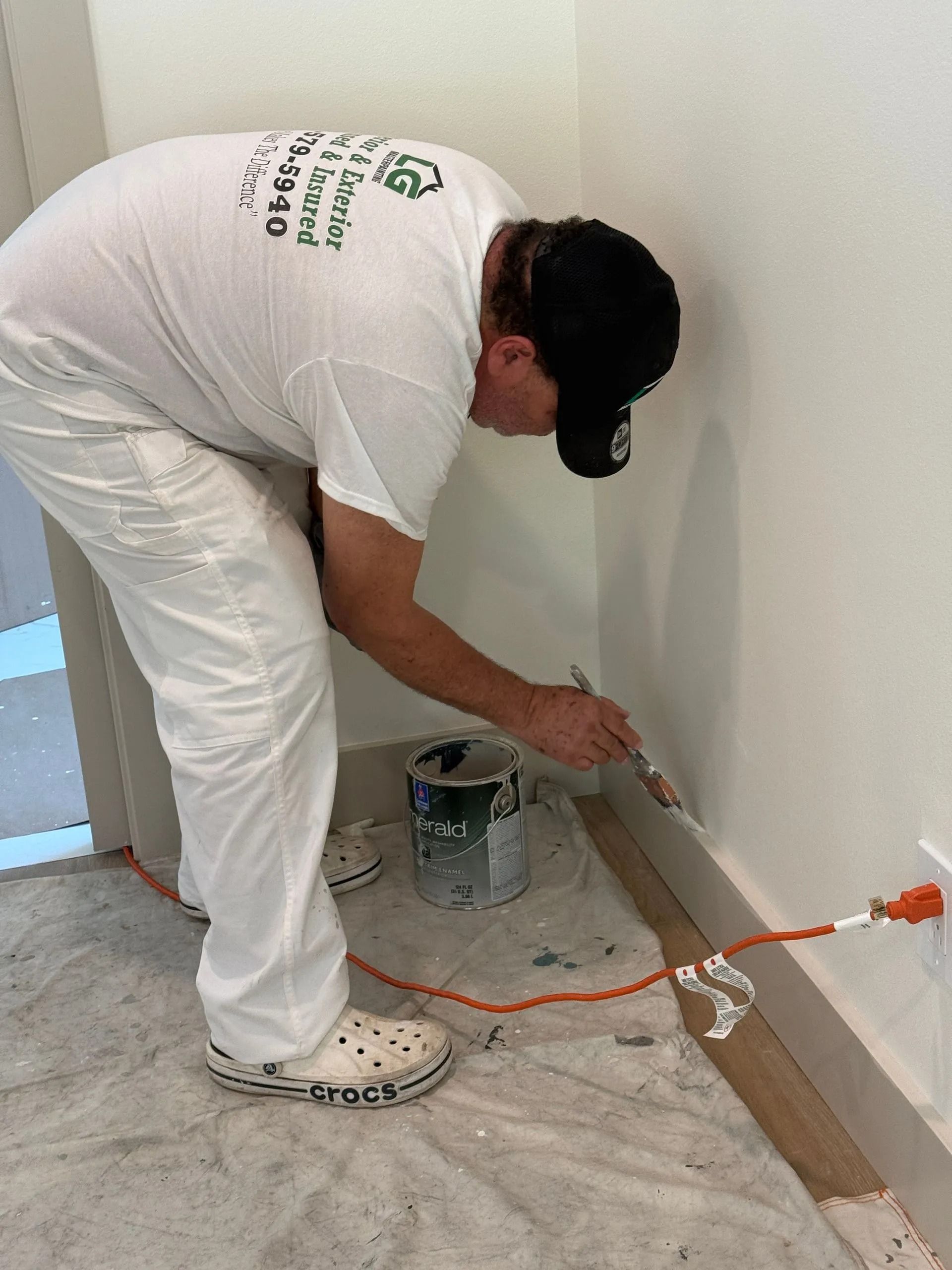 A painter in white overalls and a black cap is painting a white wall's trim with a brush.