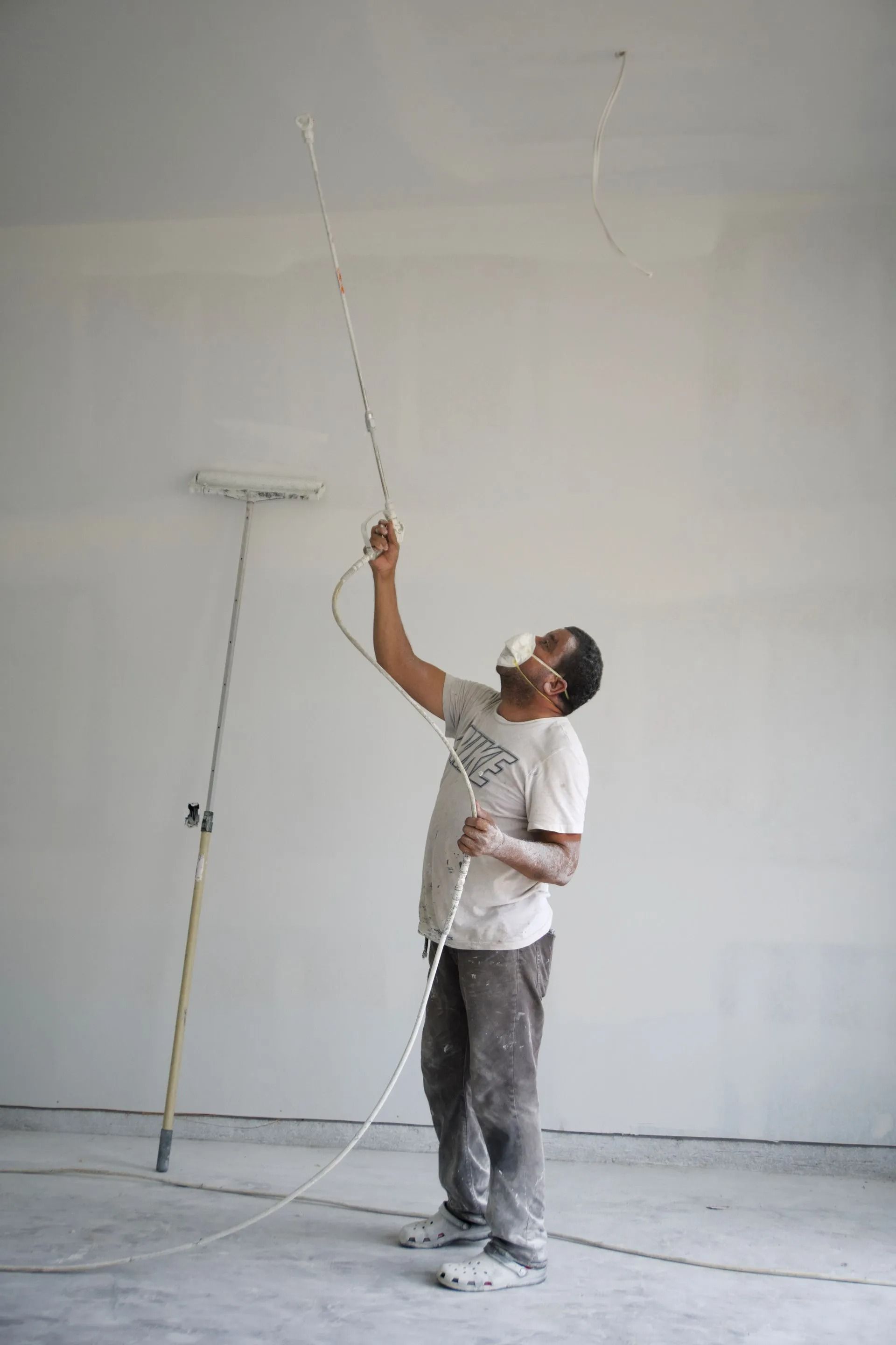 Man in a mask painting a ceiling with a spray gun in an unfinished room.