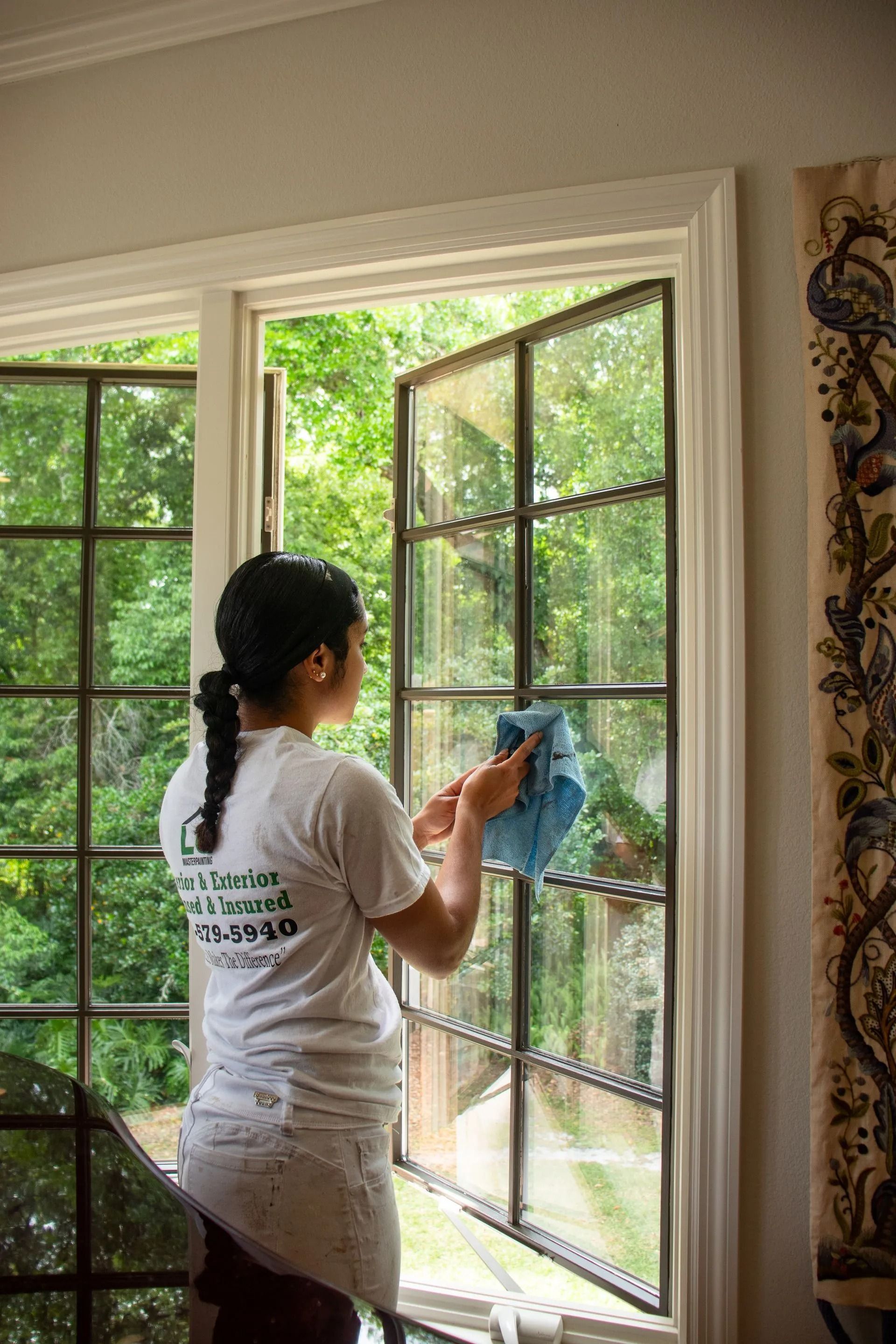 Woman cleaning a window; outdoors visible. She wears a white shirt, blue cloth in hand.