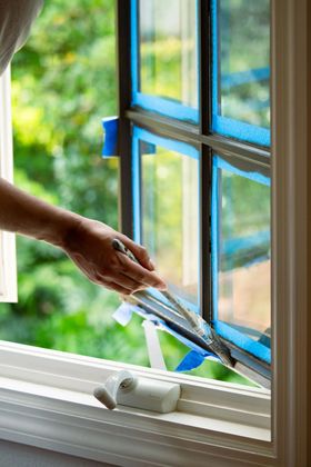 Person using a putty knife to apply caulk to a window frame, blue tape protecting the glass, outdoors.