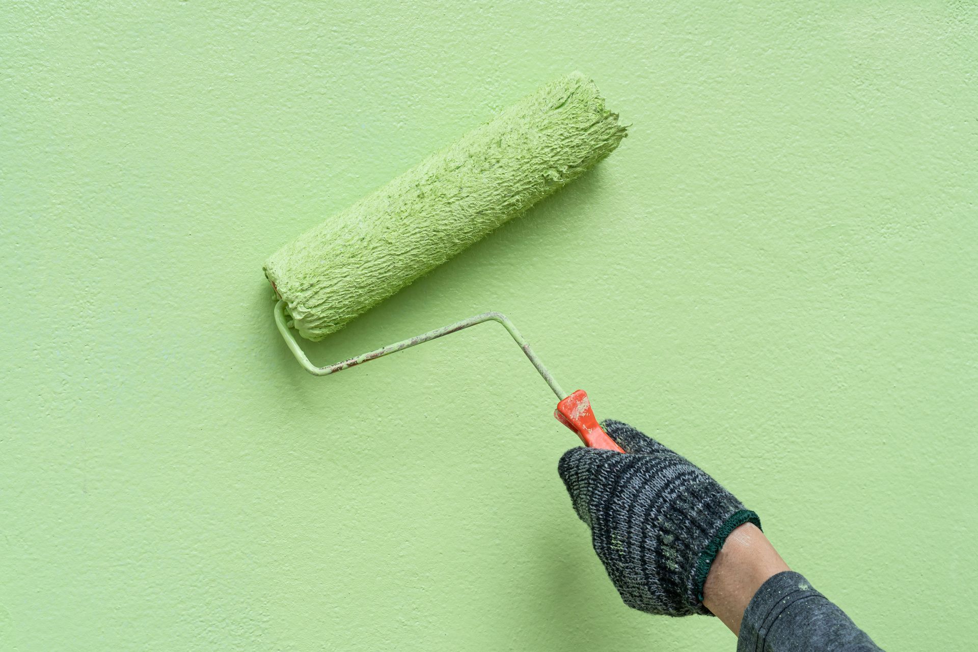 Hand wearing glove paints a green wall with a green paint roller.