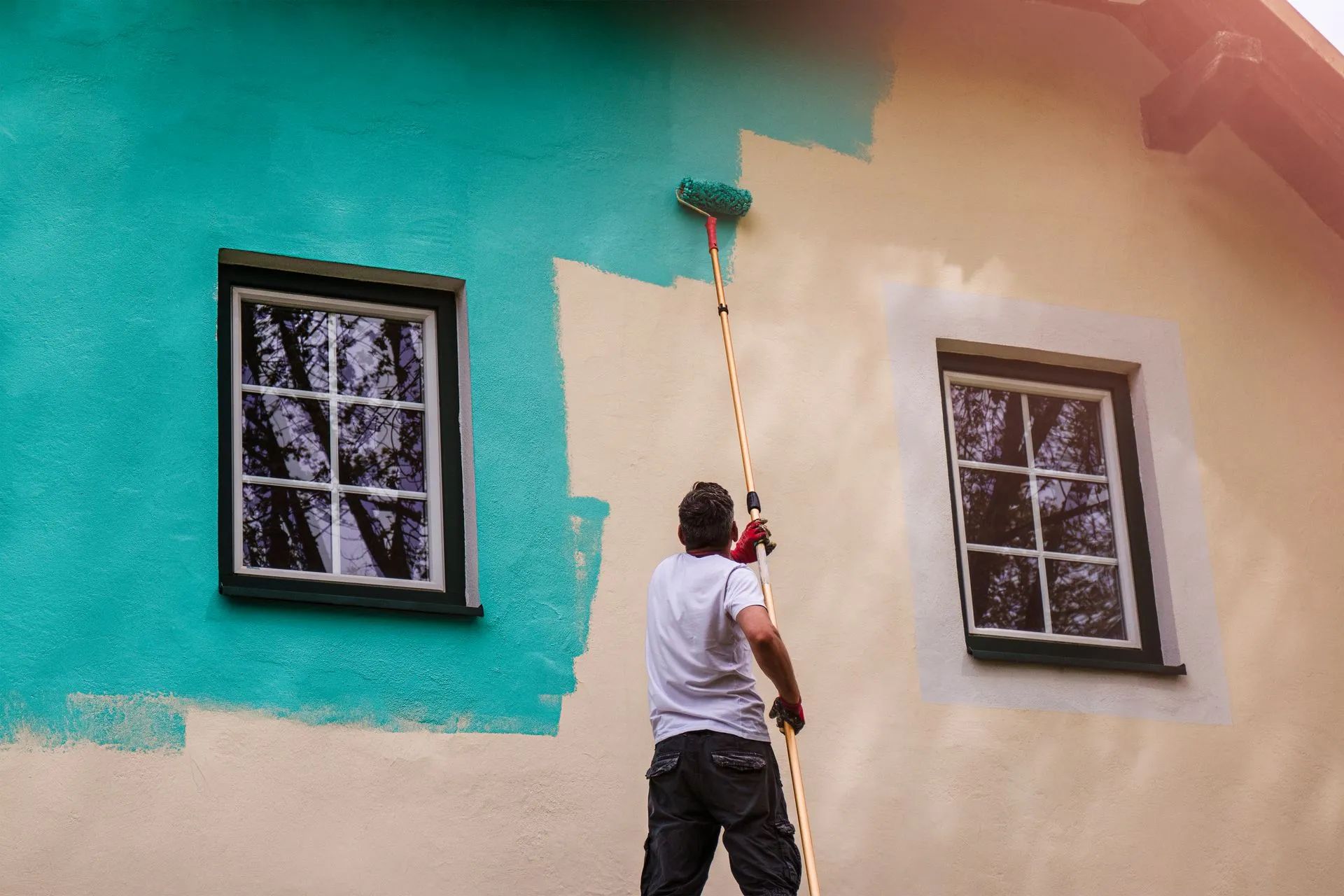 A person painting a house wall turquoise with a long-handled roller; windows on either side.