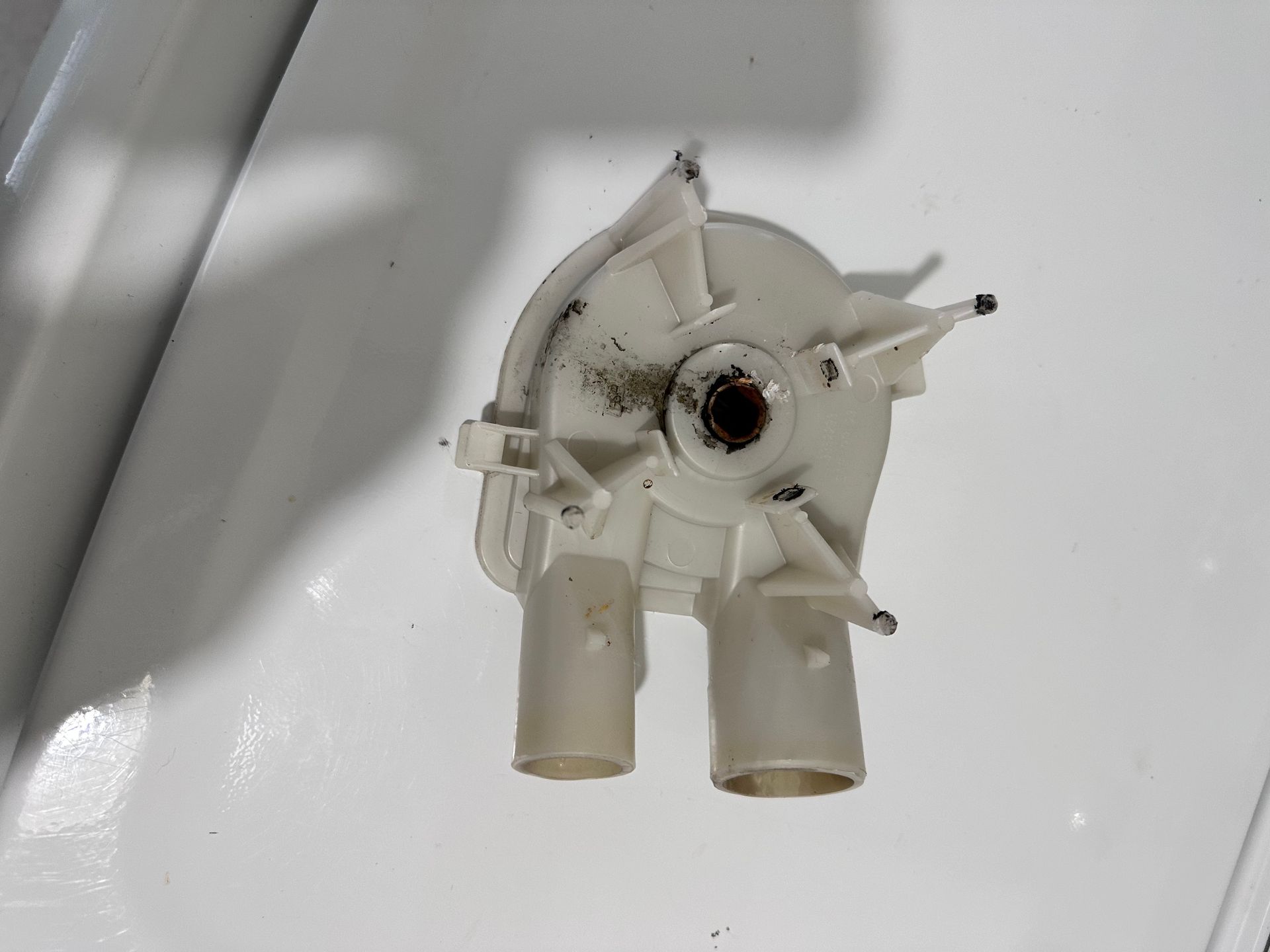 A white plastic washing machine pump, dirty with grime, featuring two spouts and a central hub.