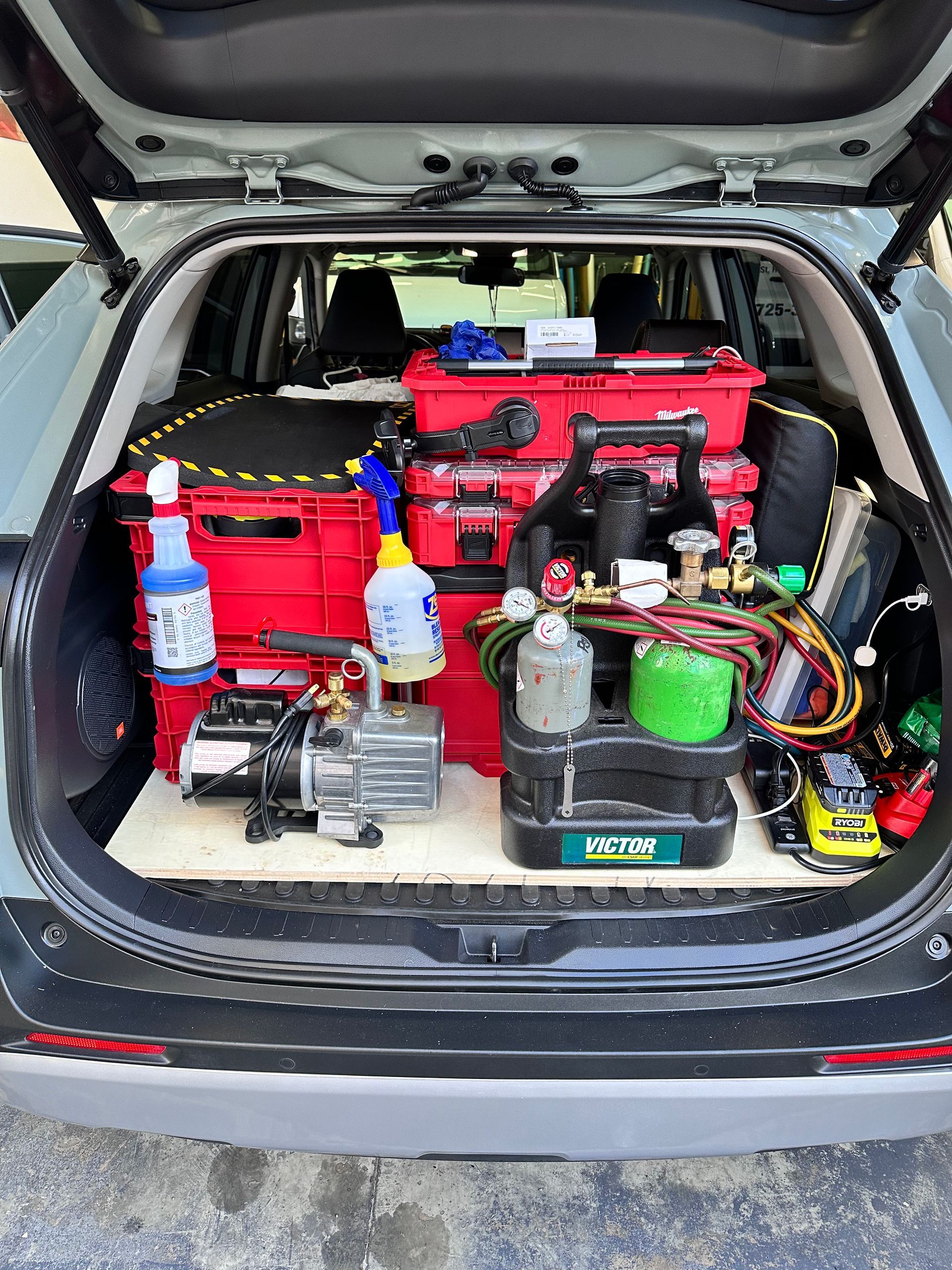 SUV trunk packed with red toolboxes, equipment, and supplies for HVAC or refrigeration service. Includes a vacuum pump, refrigerant gauges, and cleaning solutions.