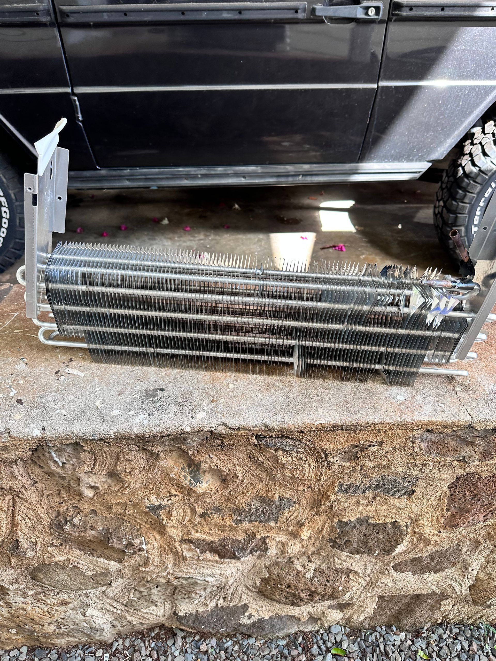 A silver car radiator lying on a weathered stone surface, possibly for repair or replacement. It is positioned in front of a black vehicle.
