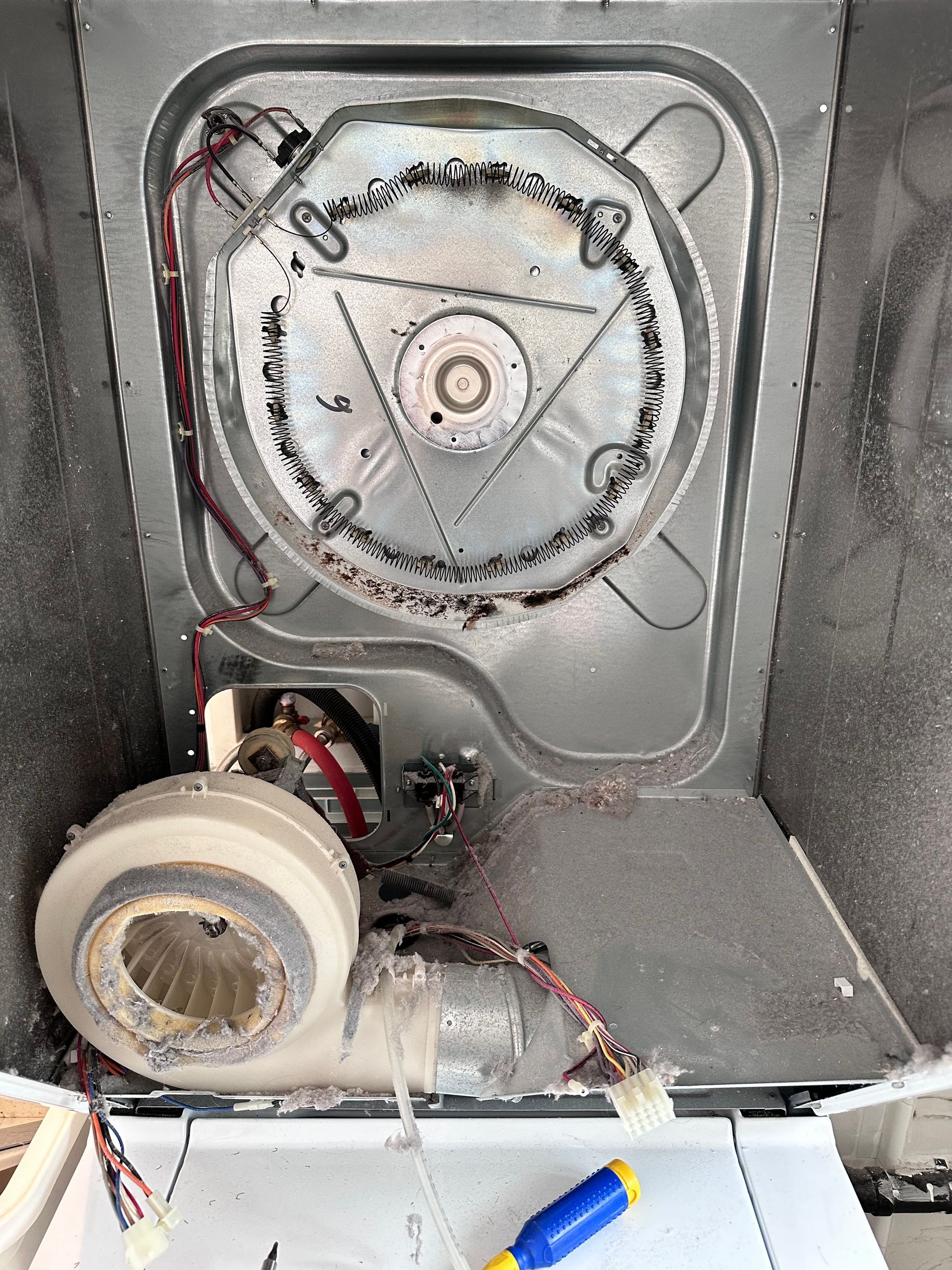 Interior of a dryer, showing the heating element, fan, and lint accumulation. Grey metal with wires and a blue and yellow tool.