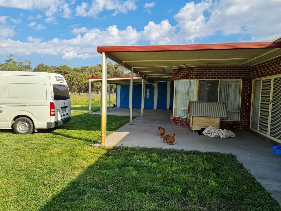 A white van parked beside a brick house with a covered patio. Two small dogs on the patio.