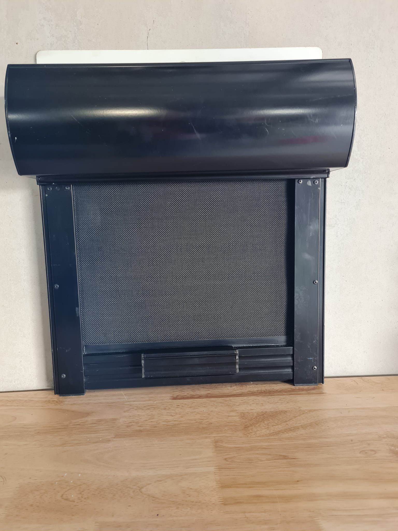 Black roller blind, mesh screen visible, installed against a light-colored wall and wooden floor.