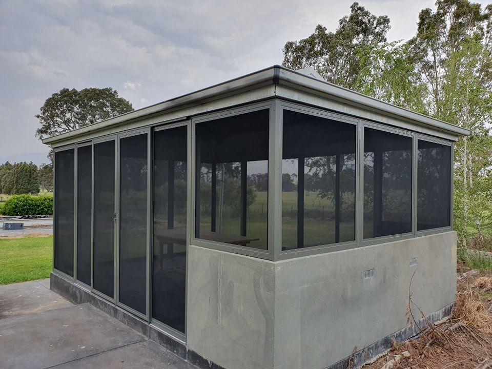 A screened-in gazebo with concrete walls, on a gray day, overlooking a grassy area.