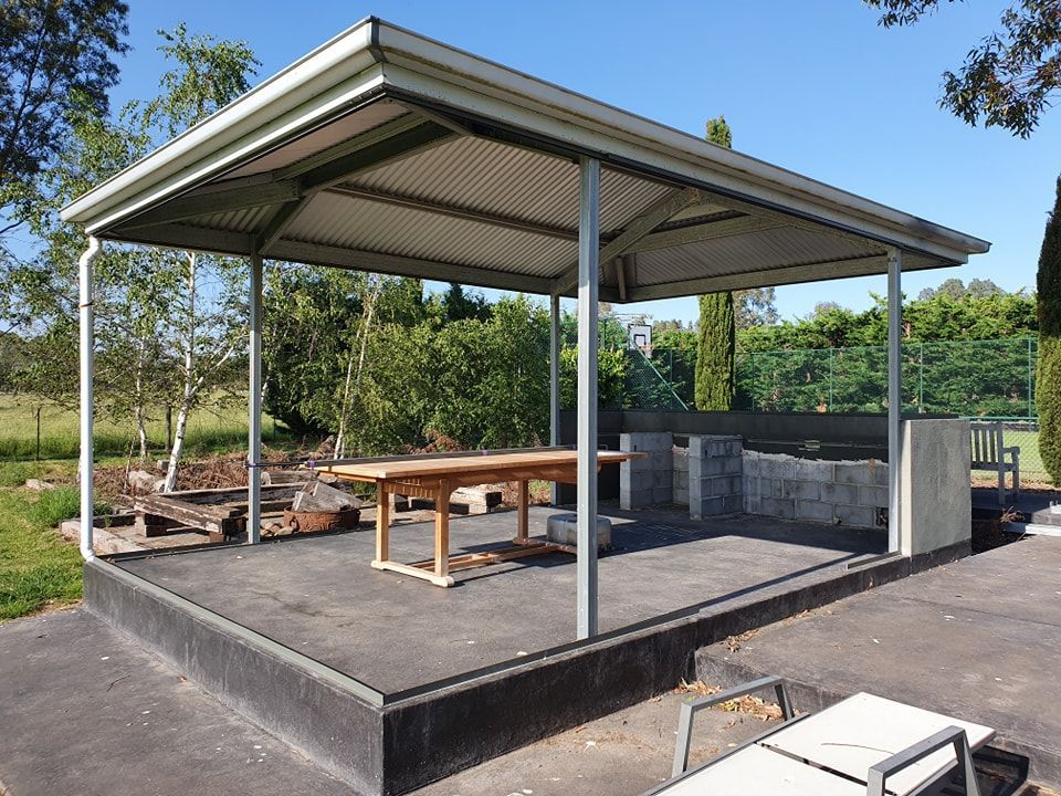 Outdoor shelter with a table and barbecue grill, trees in the background, sunny day.