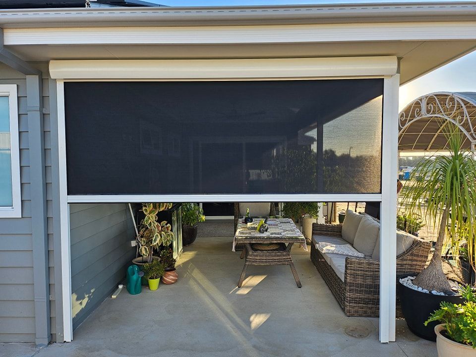 Outdoor patio with a partially lowered black roller shade, furniture, and plants.