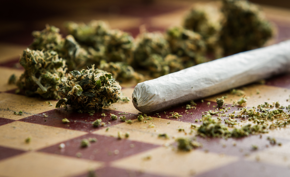 A rolled cannabis joint rests on a checkerboard surface alongside several dried cannabis buds.