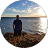 Person standing on a rocky shore facing a calm sea at sunset