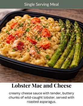 A container of lobster mac and cheese with roasted asparagus on a wooden surface, topped with breadcrumbs and herbs.