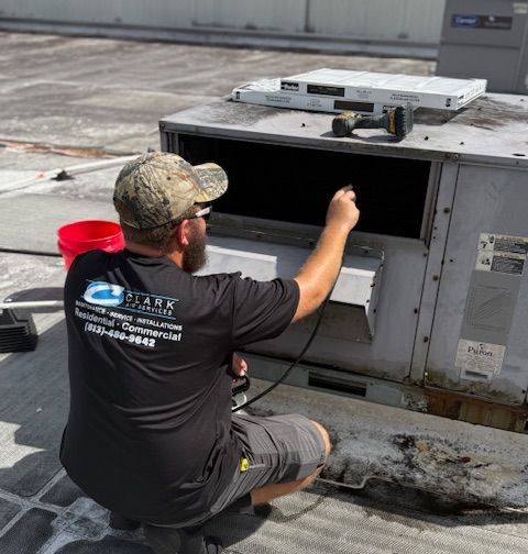 HVAC technician inspecting a rooftop unit. He wears a hat and Clark Company shirt, kneeling near the equipment.