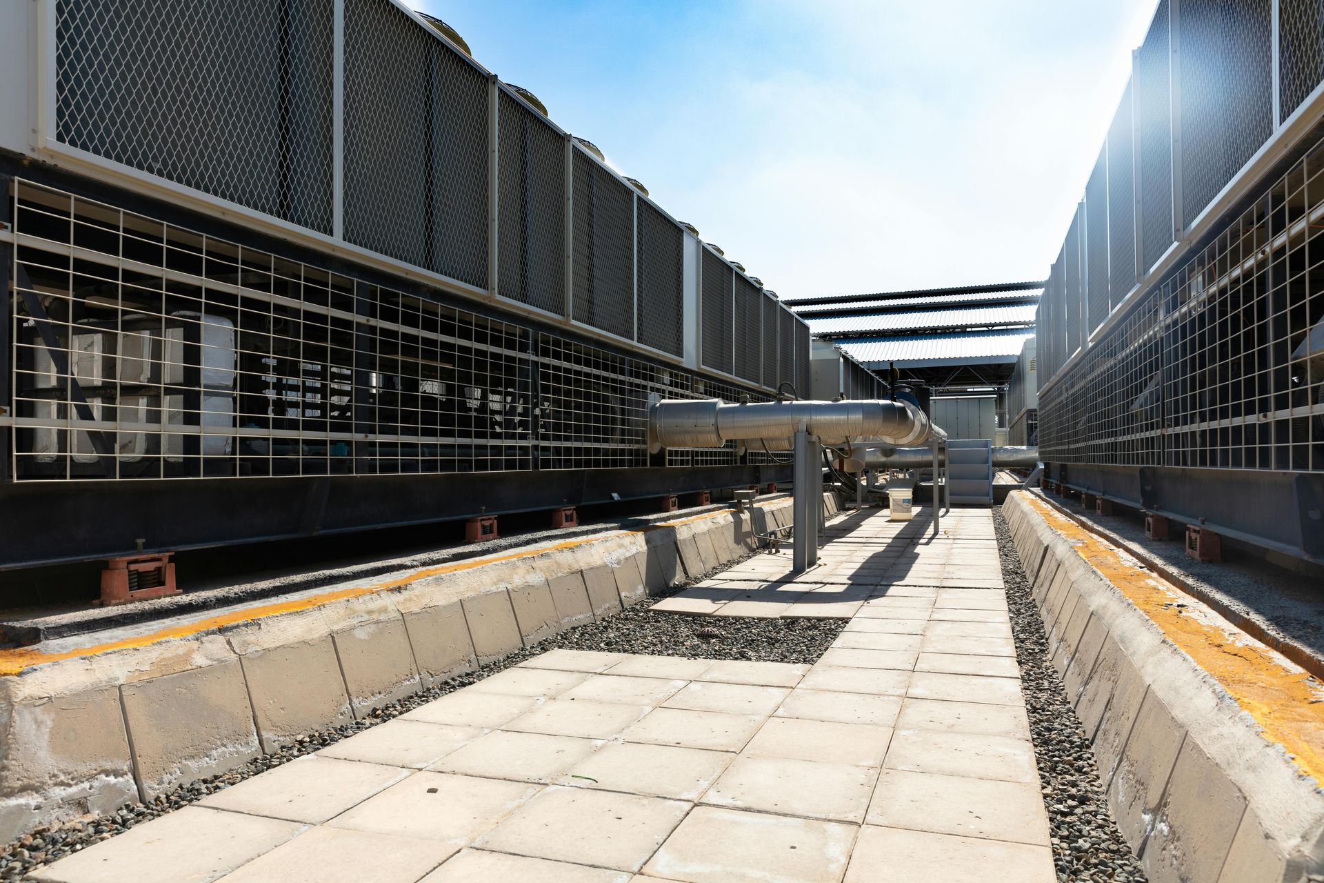 Large industrial air conditioning units on a rooftop. Bright sunlight, gray metal, concrete walkway.