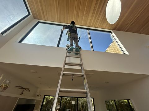 A person climbs an extension ladder inside a room with high ceilings to clean a large window.