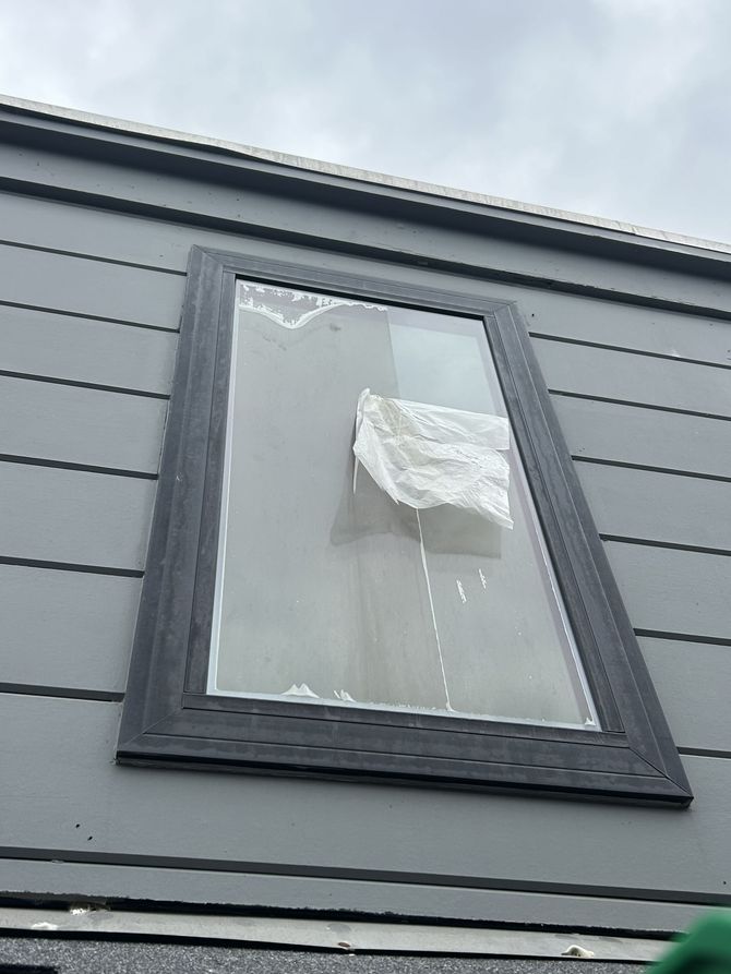 A person wearing yellow gloves kneels on a roof, using a small tool to clear debris from a gutter.