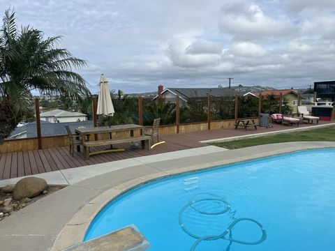 A bright blue swimming pool sits next to a wooden deck with patio furniture, overlooking a neighborhood under cloudy skies.