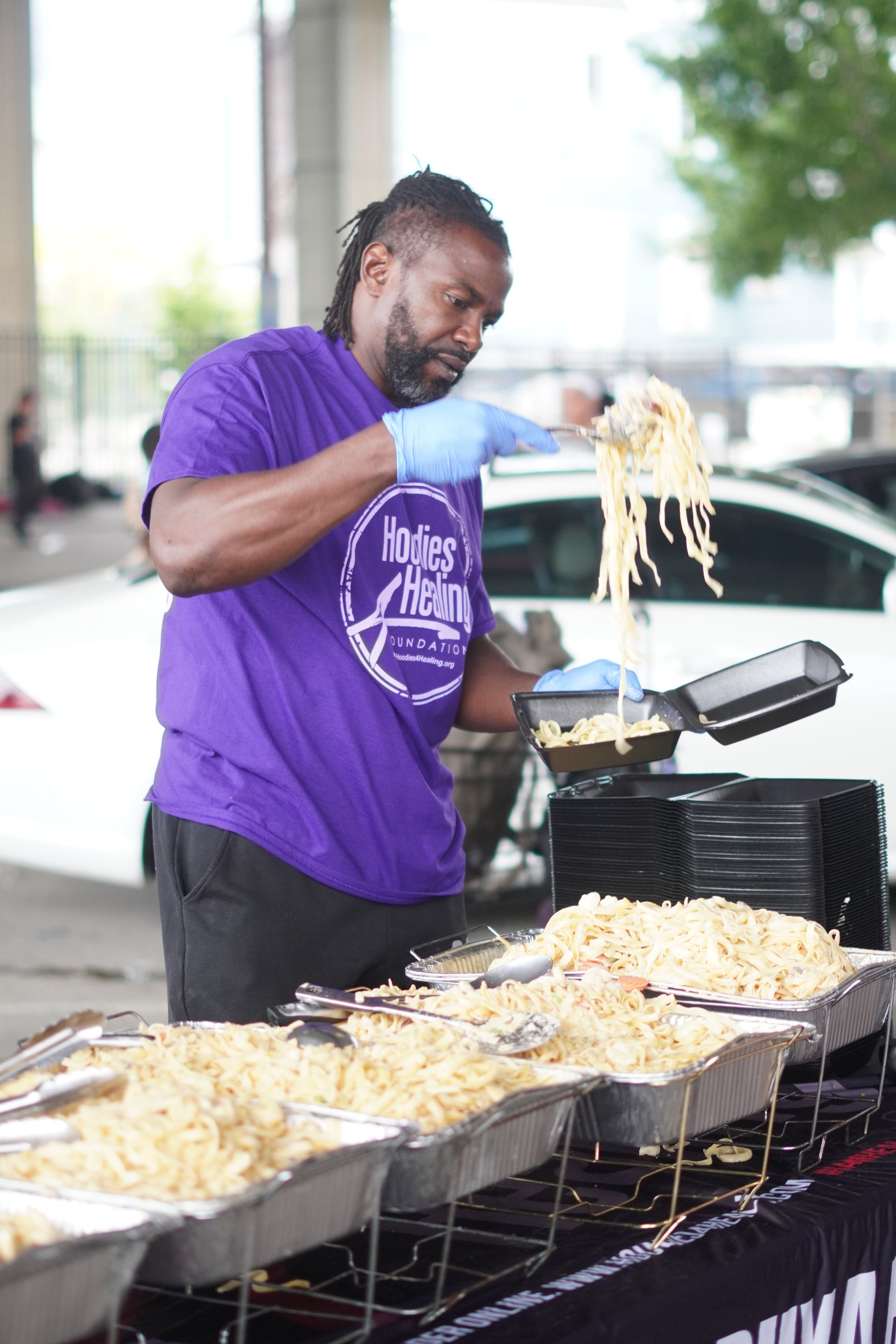 A man in a purple shirt is cooking noodles in a frying pan.