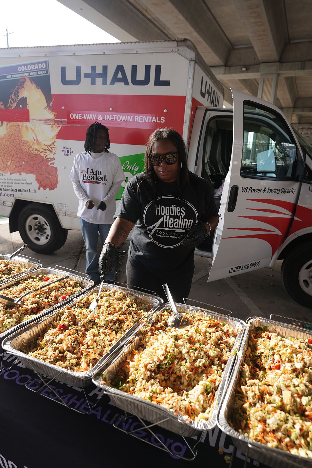 A woman is standing in front of a uhaul truck filled with trays of food.