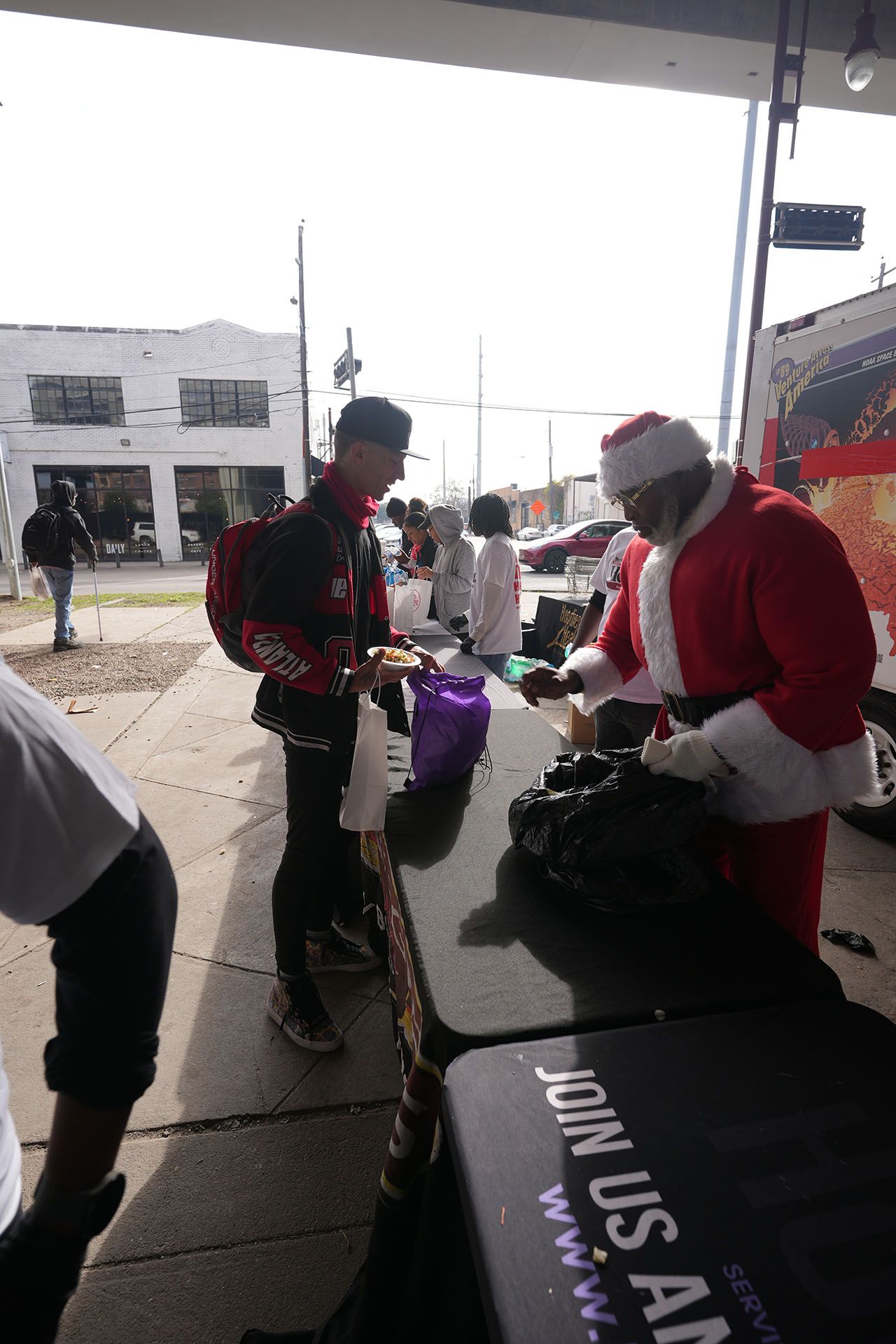 A man dressed as santa claus is standing in front of a table that says join us all