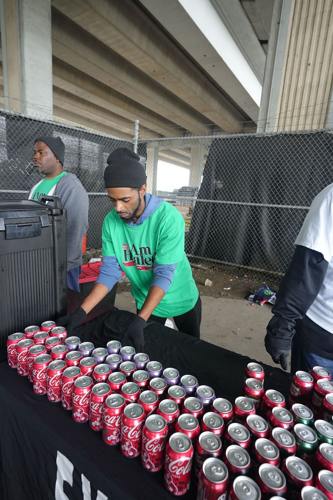 A man in a green shirt is standing in front of a table filled with coca cola cans.