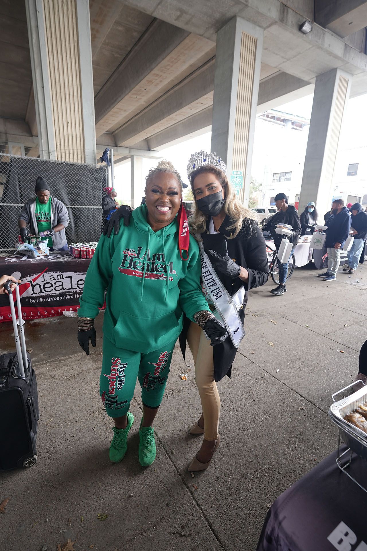 Two women are standing next to each other on a sidewalk under a bridge.