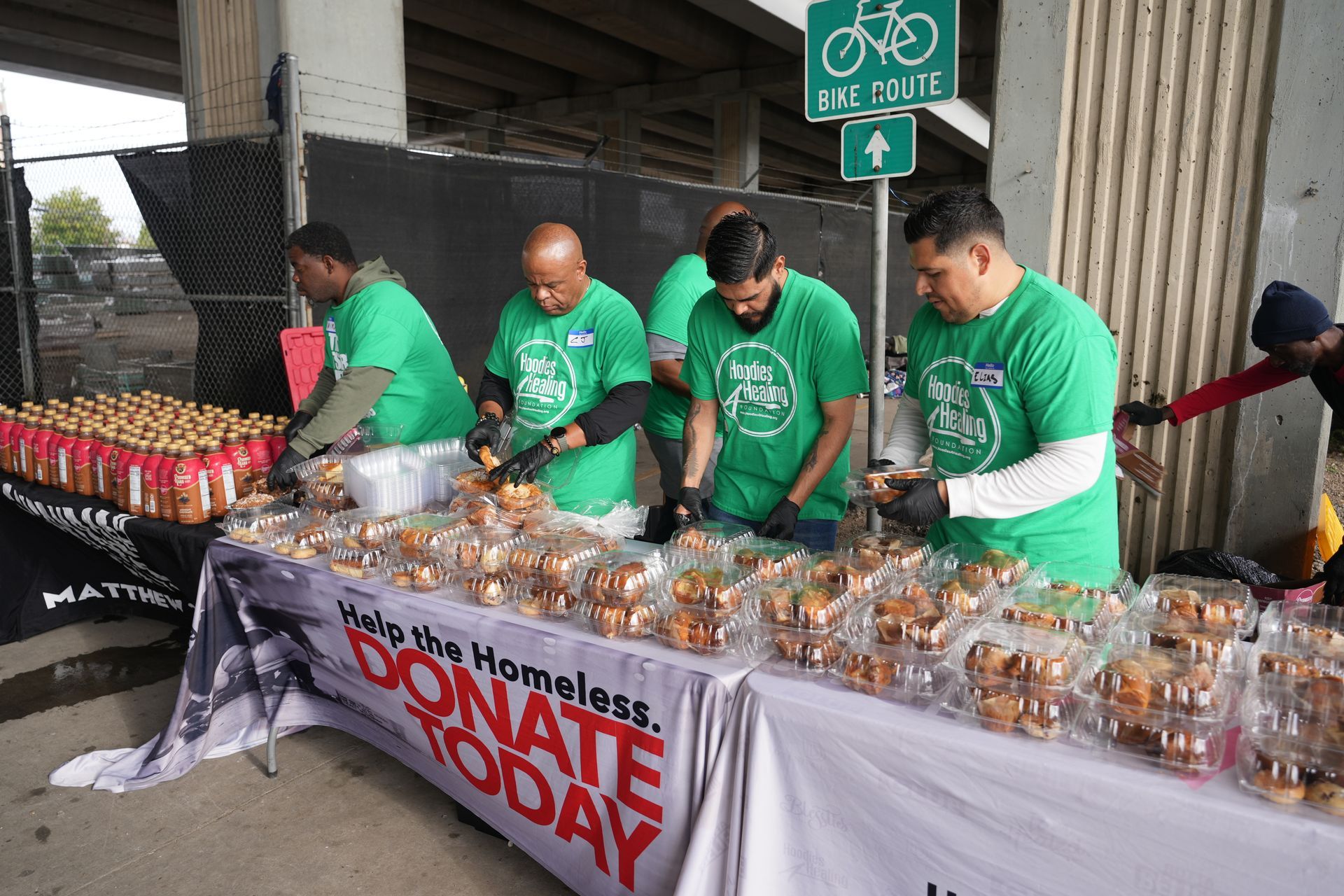 A group of men in green shirts are standing around a table filled with food.