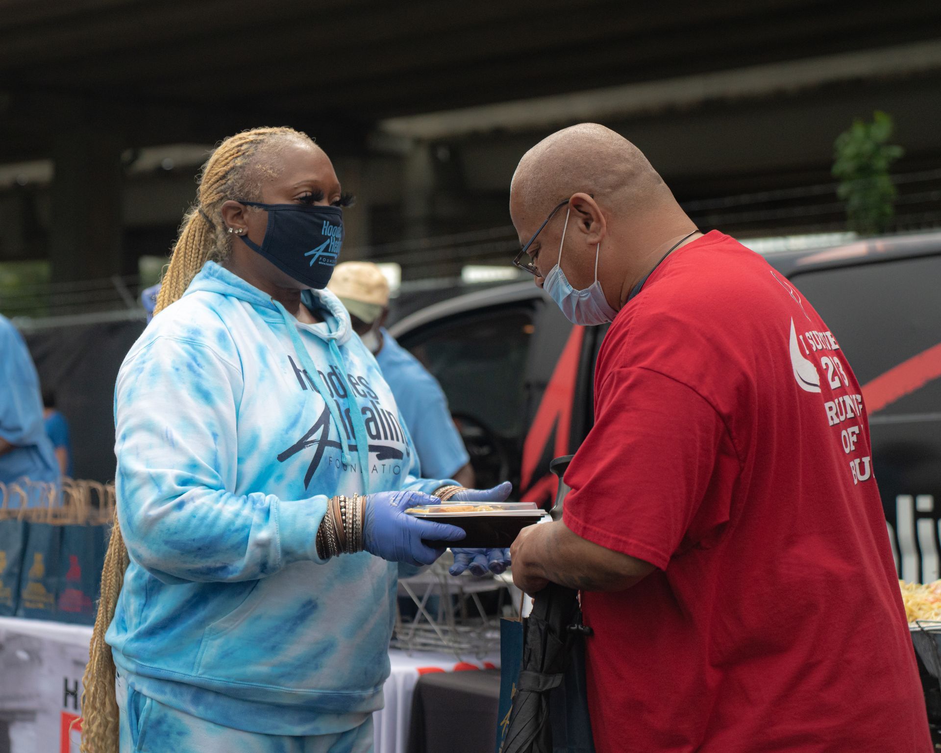 A man in a red shirt is giving a plate of food to a woman in a blue tie dye sweatshirt.