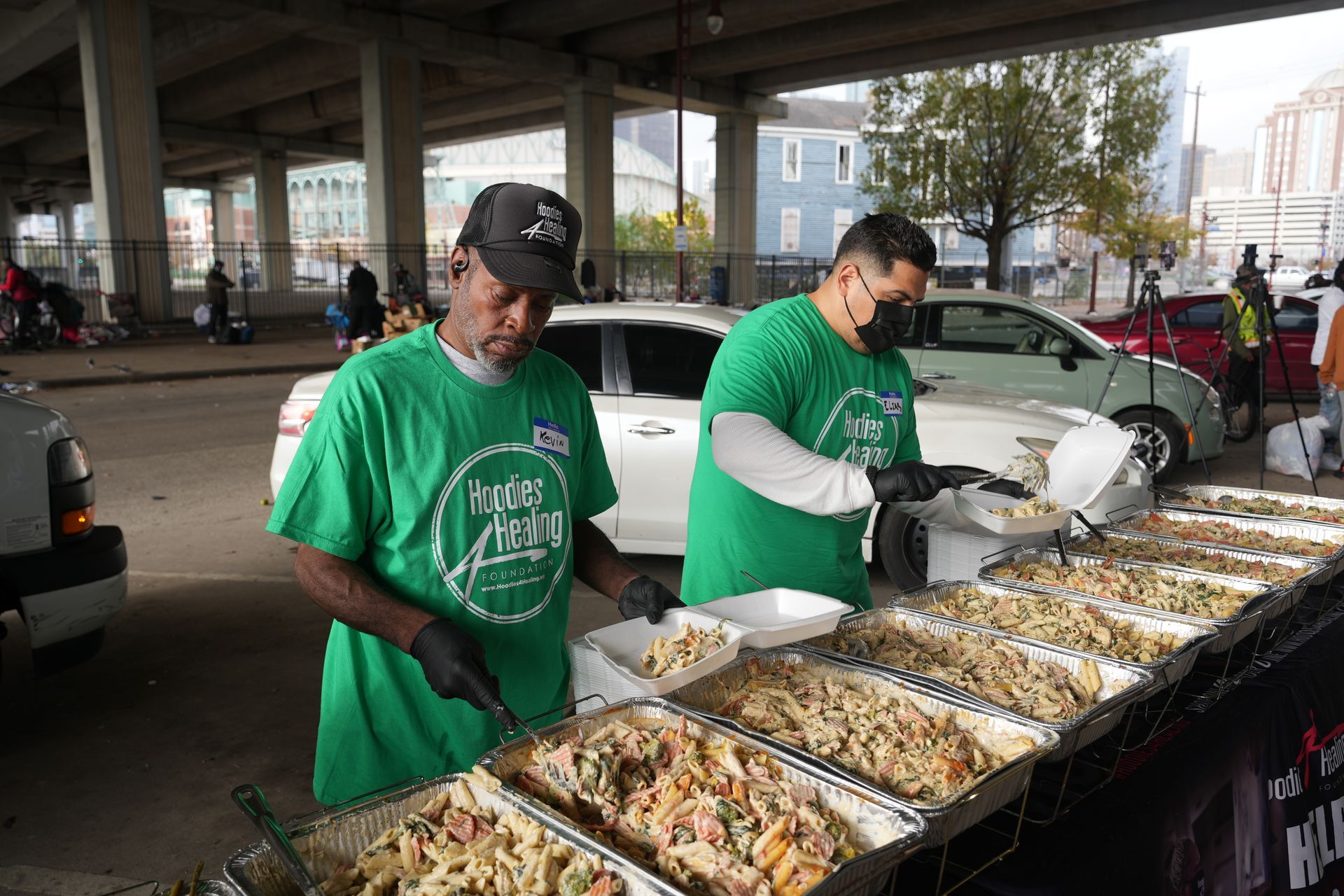 Two men in green shirts are preparing food in a parking lot.