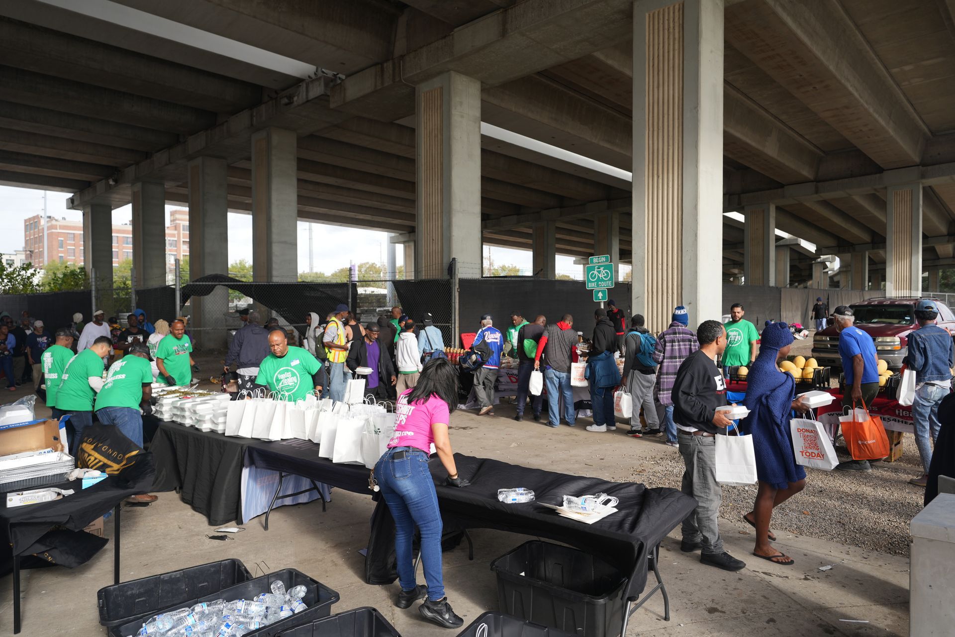 A group of people are standing around tables under a bridge.