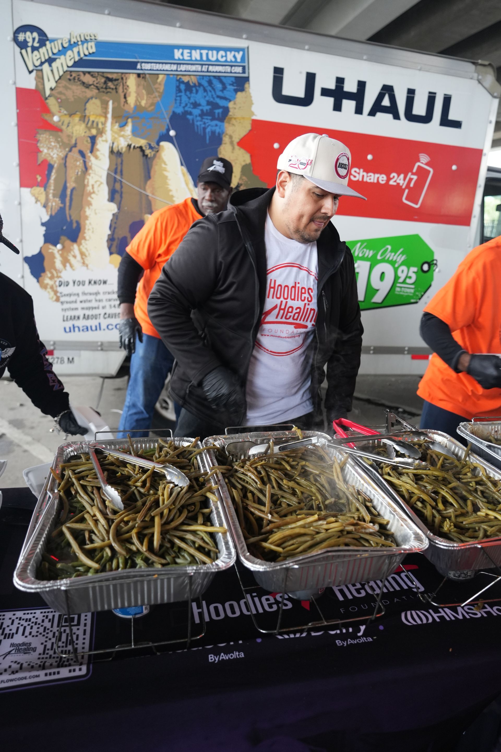 A man is standing at a table with trays of food in front of a u-haul truck.