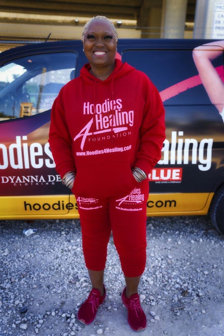 A woman in a red hoodie is standing in front of a van that says hoodies 4 healing