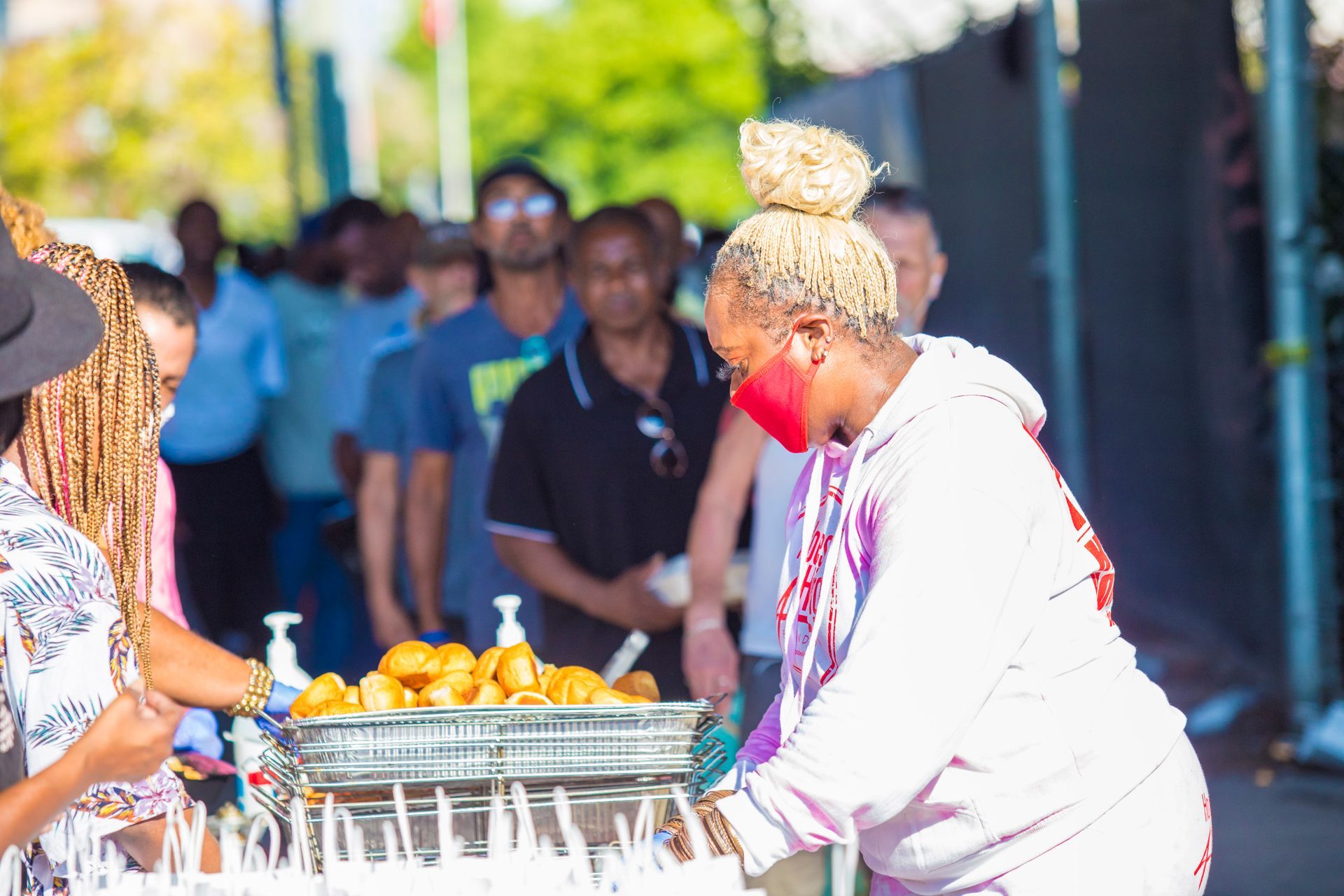 A woman wearing a red mask is serving food to a crowd of people.