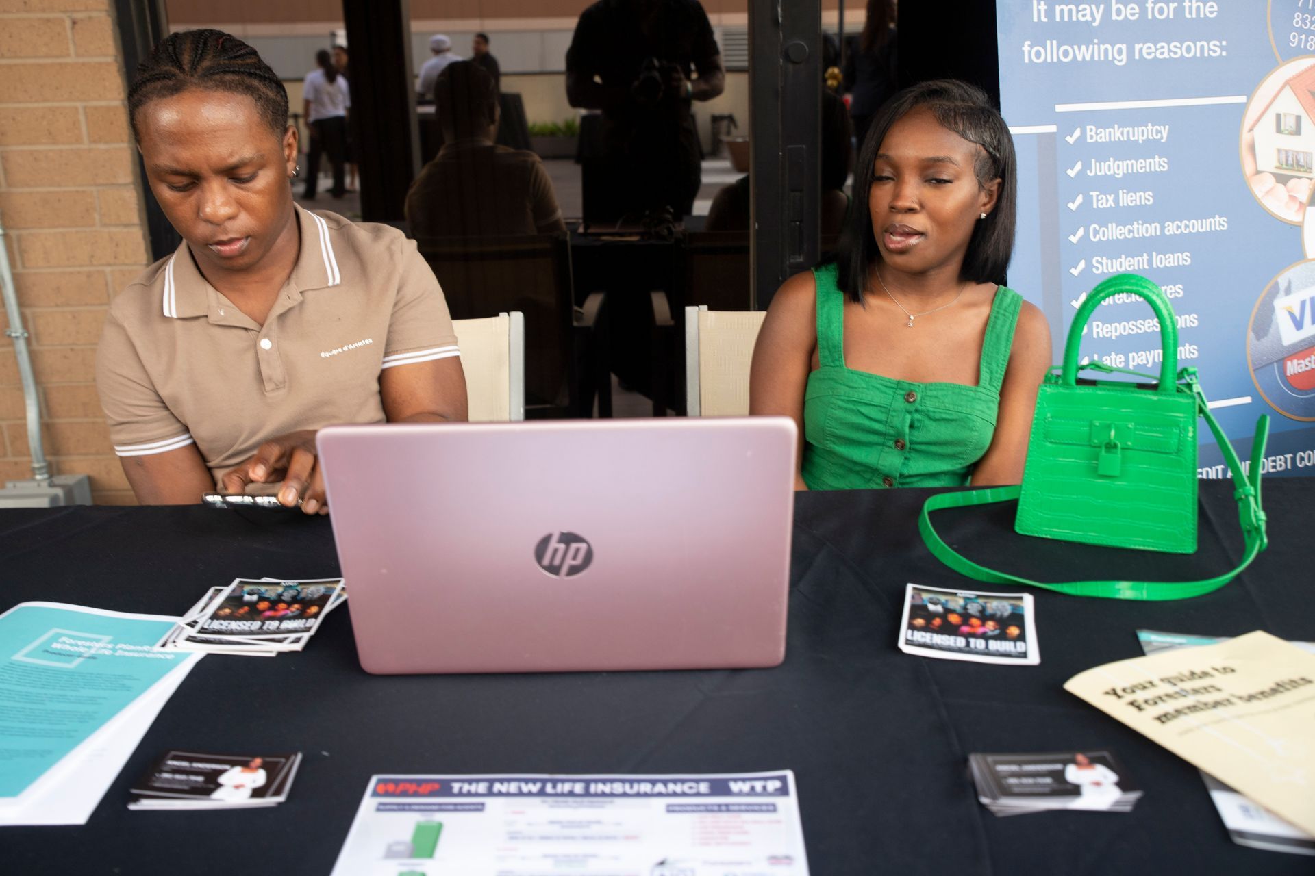 Two women are sitting at a table with a laptop and a green purse.