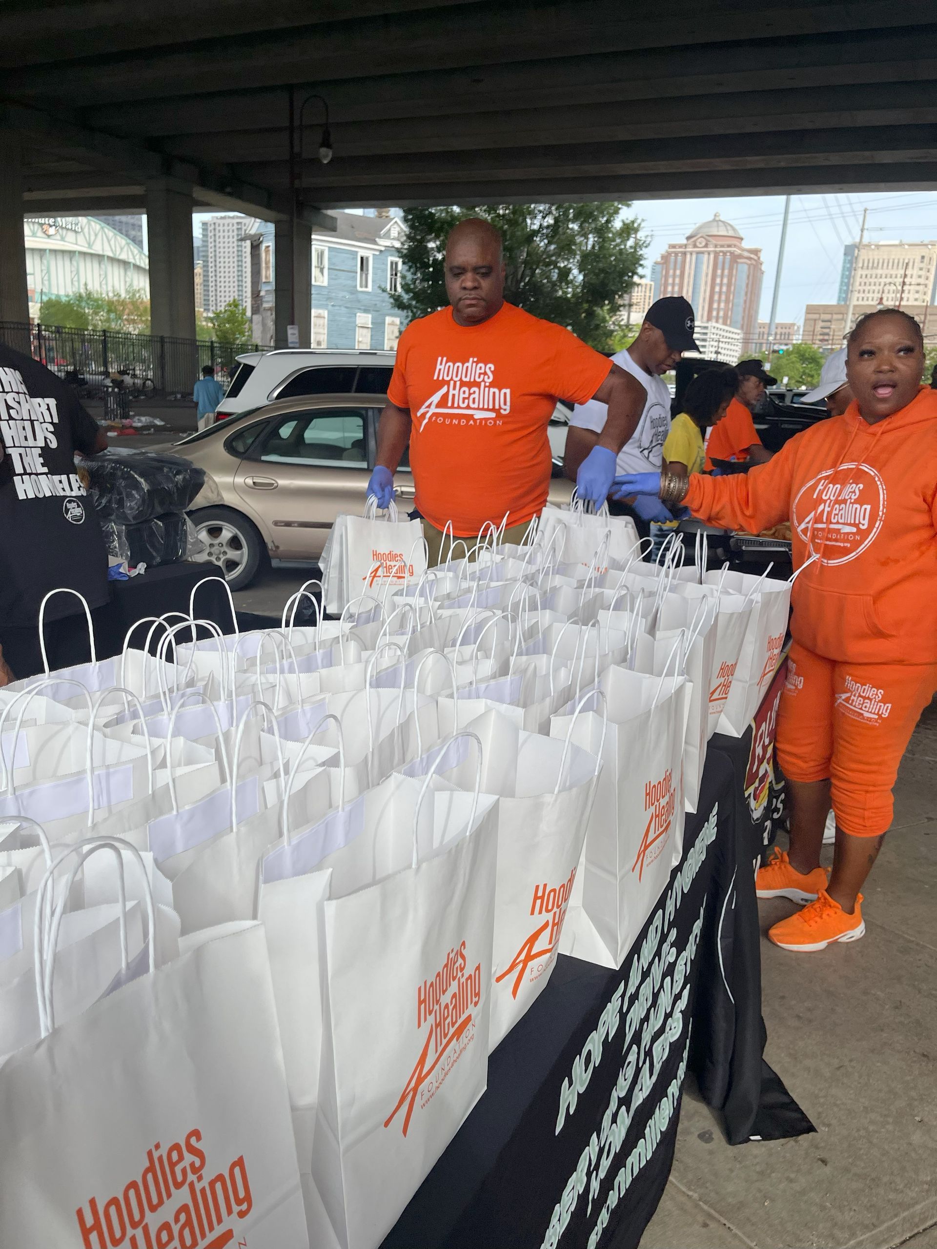 A man in an orange shirt is standing next to a table full of bags that say hoodies being