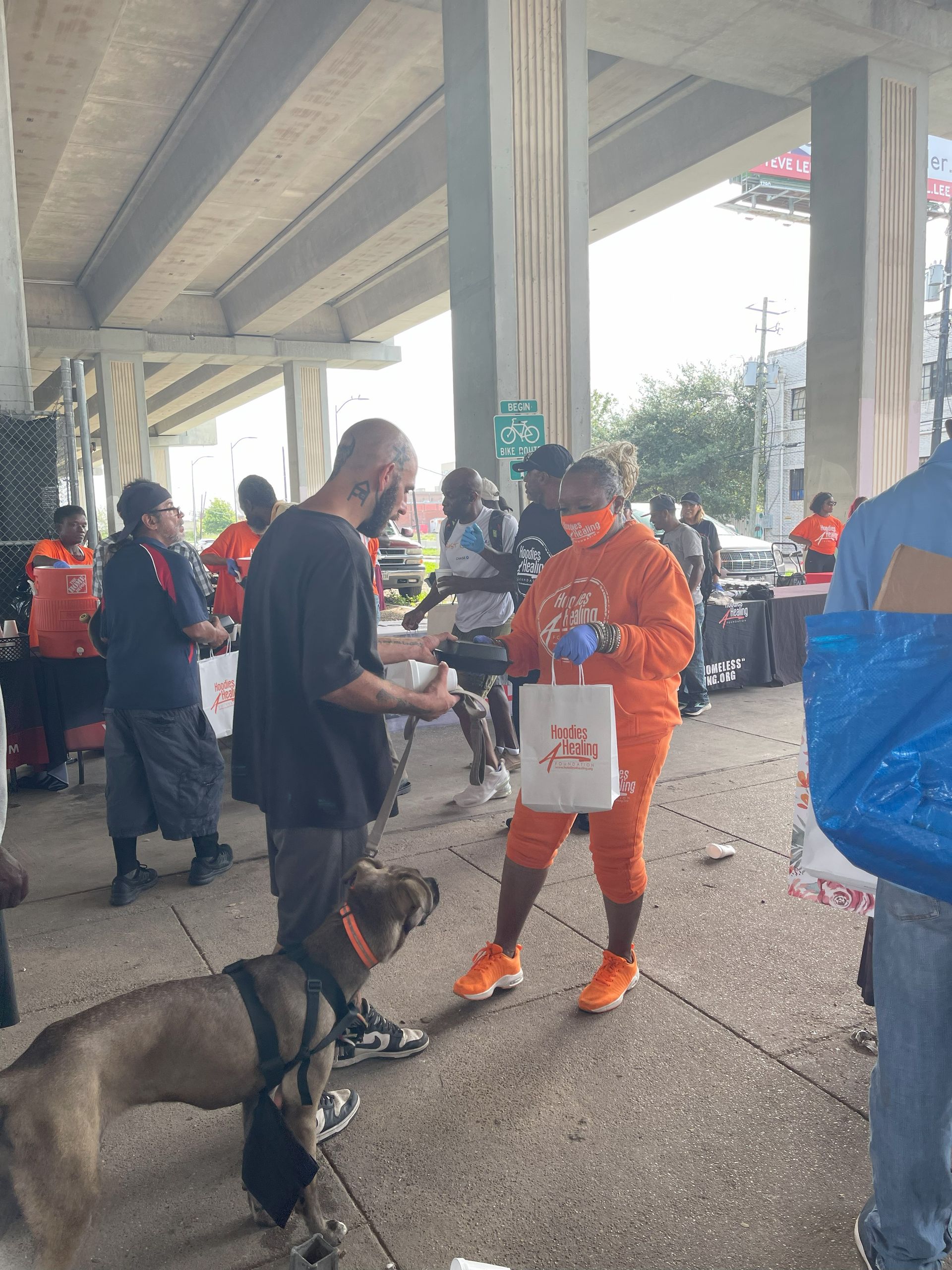 A group of people are standing on a sidewalk under a bridge.