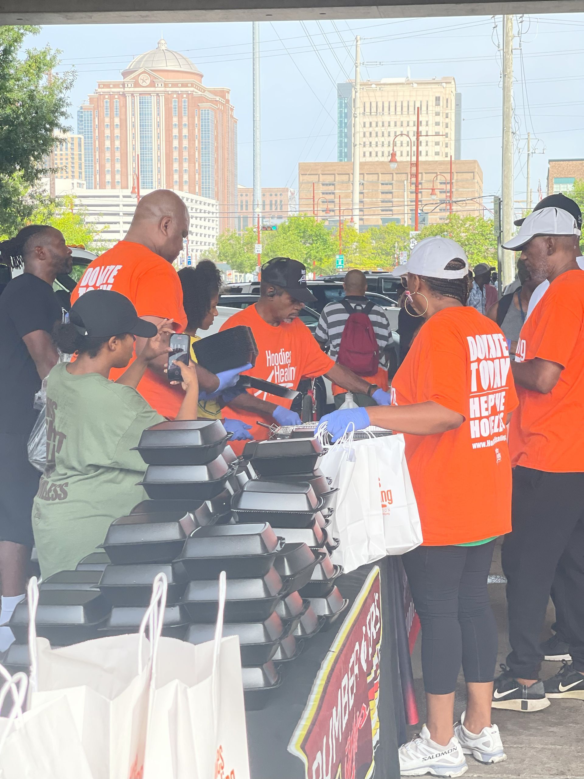 A group of people are standing around a table with boxes on it and one of them is wearing an orange shirt that says biker tuff