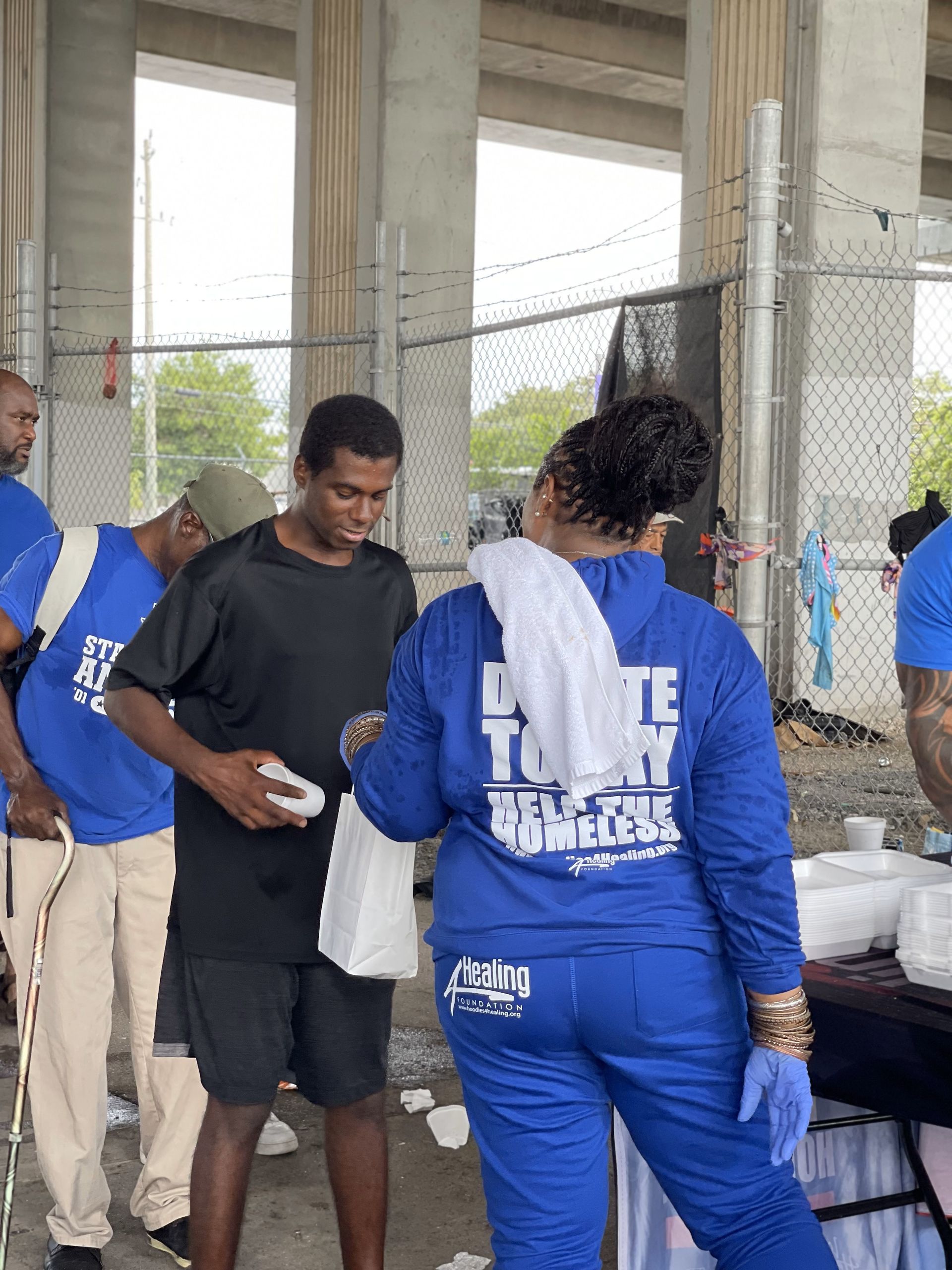 A group of people are standing around a table with a woman wearing a blue shirt that says i love to help others.