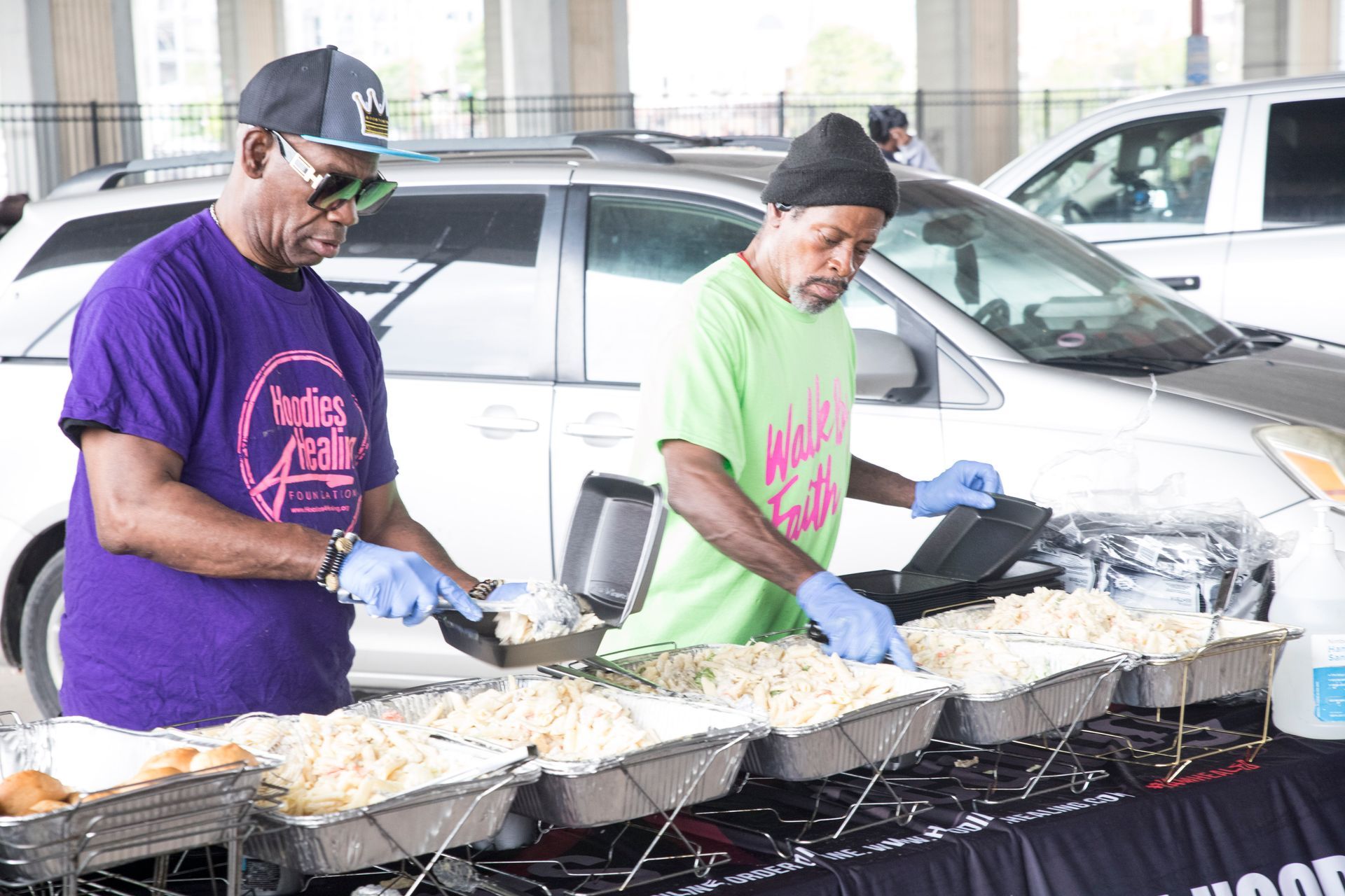 Two men are cooking food in front of a car in a parking lot.
