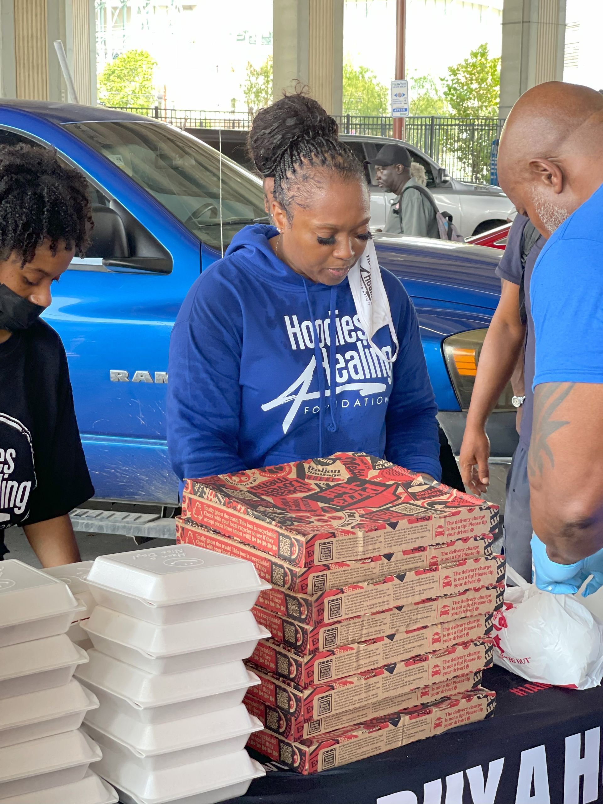 A woman in a blue hoodie is standing next to a stack of pizza boxes.
