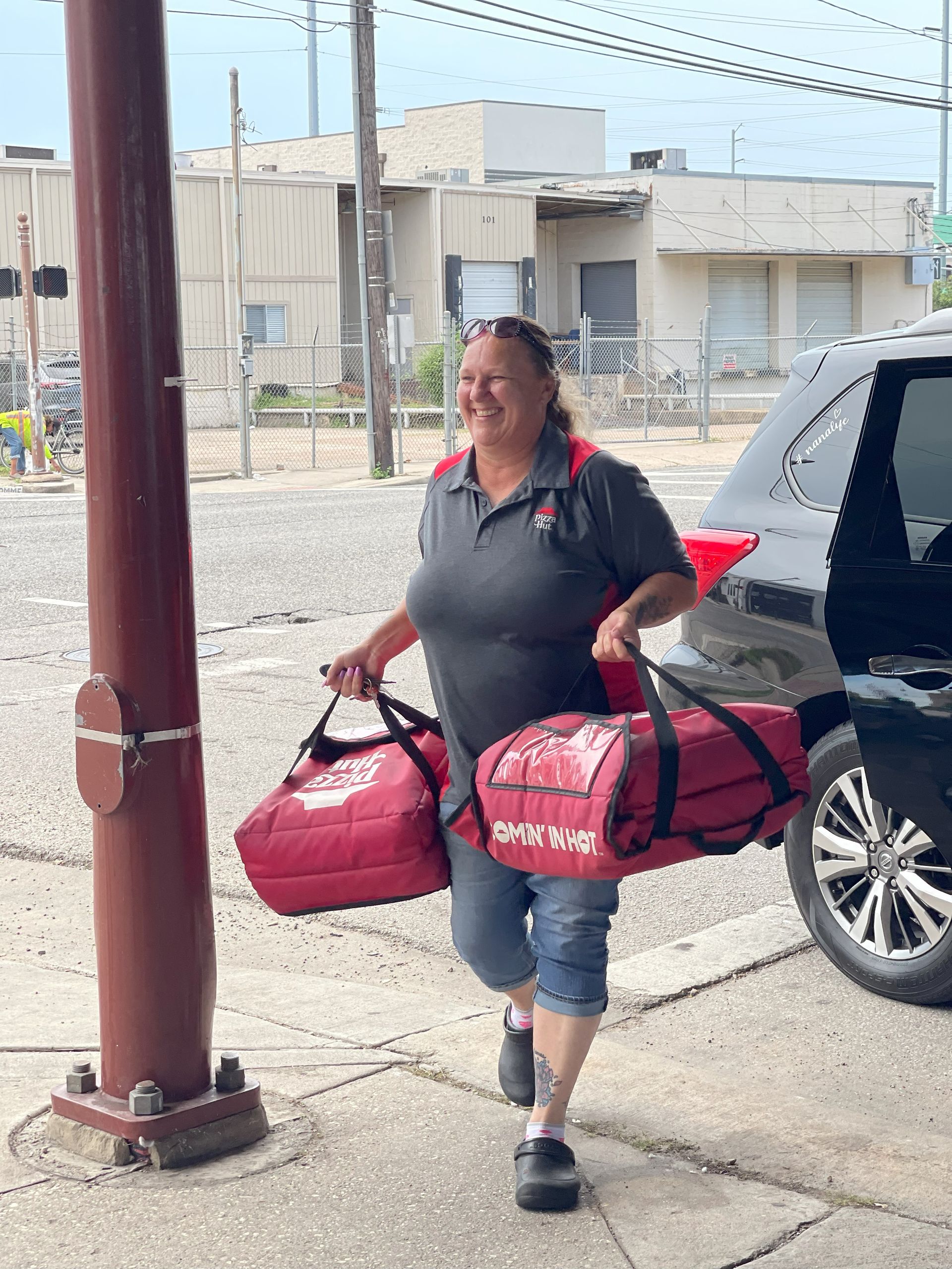 A woman is carrying two bags of pizza next to a car.