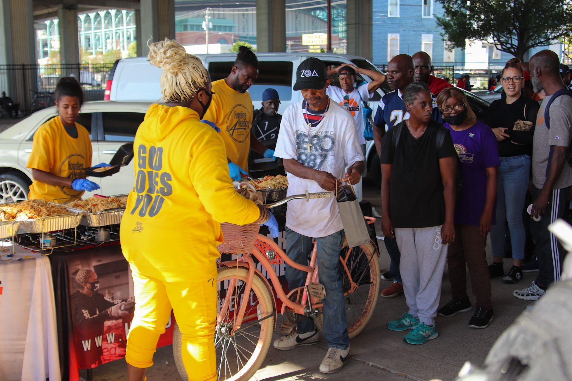 A group of people are standing around a table with a woman wearing a yellow hoodie that says go des