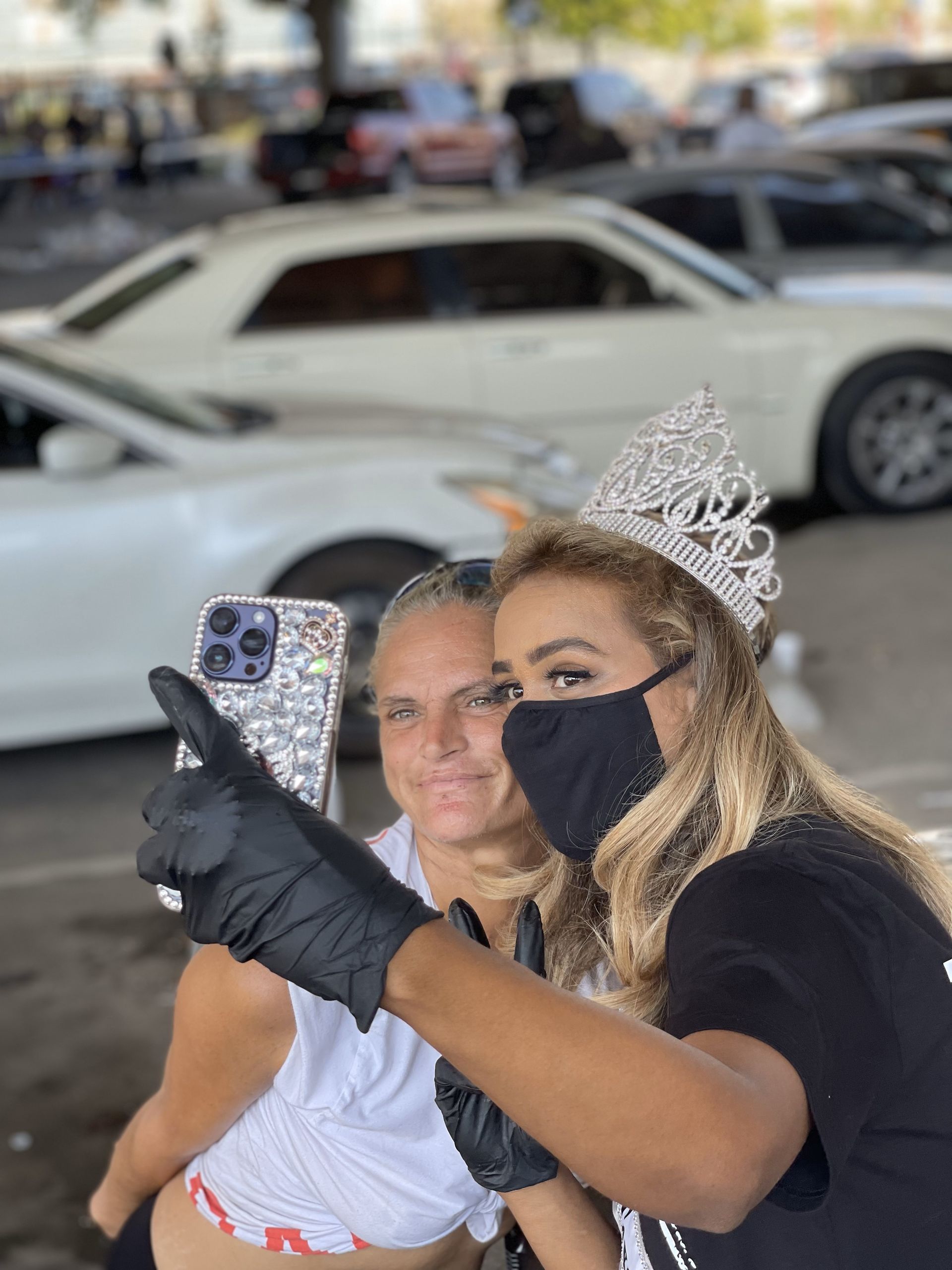 Two women wearing masks and tiaras are taking a selfie in a parking lot.