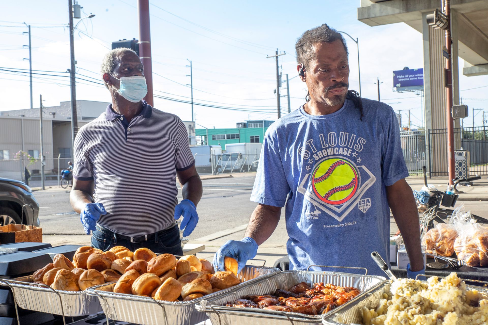 Two men wearing masks and gloves are standing in front of a table of food.