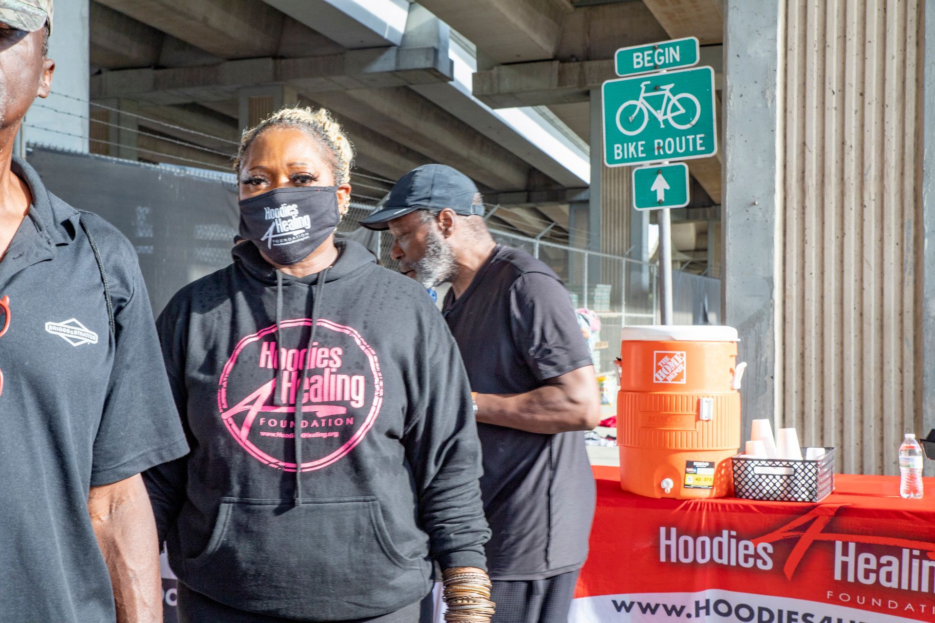 A woman wearing a mask is standing in front of a table with hoodies on it.
